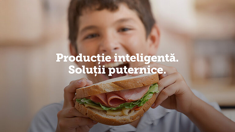 Băiat zâmbind și ținând un sandwich mare cu șuncă, salată și brânză; text suprapus: ‘Producție inteligentă. Soluții puternice.’