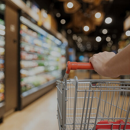 Ein Einkaufswagen wird durch einen Supermarkt auf den Wurstbereich zugeschoben. Von der einkaufenden Person sieht man nur einen Teil des Arms. 