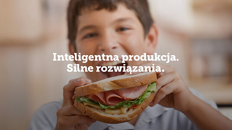 Chłopiec trzyma dużą kanapkę z szynką i świeżymi warzywami; na środku widoczny jest napis ‘Inteligentna produkcja. Silne rozwiązania.