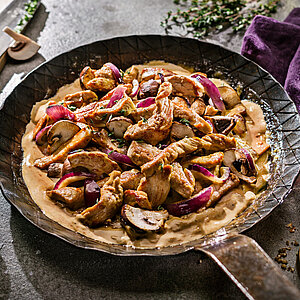 Pfanne mit gebratenen Fleischstreifen, roten Zwiebeln und Pilzen in einer cremigen Sauce, rustikal angerichtet.