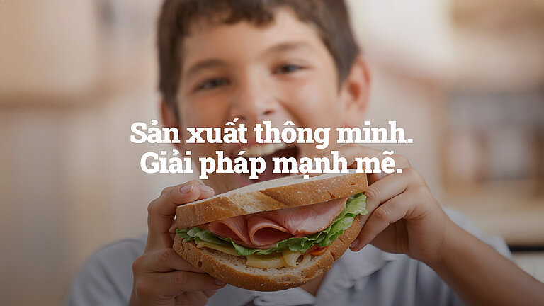 Đứa trẻ cười tươi cầm chiếc sandwich lớn với thịt nguội và rau xà lách; văn bản trên hình ghi: ‘Sản xuất thông minh. Giải pháp mạnh mẽ.’
