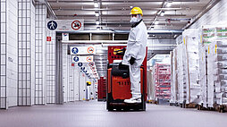 Ein Mitarbeiter von Gustoland in weißem Schutzanzug, Mundschutz und gelbem Schutzhelm steht in einem langen Lagerhallenflur hinten auf einem Hubwagen. Am rechten Rand des Flures stehen fertig verpackte Produkte auf Paletten. Der Flur ist weiß gefliest.