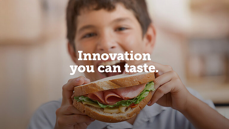 Boy eating a large sandwich with ham, lettuce and cheese while smiling.