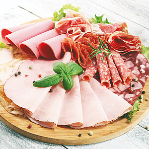 Vielfältige Aufschnittplatte mit verschiedenen Schinken-, Wurst- und Salamisorten, garniert mit Kräutern und Pfefferkörnern auf einem runden Holzbrett.